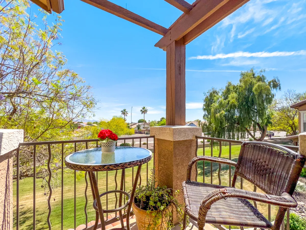 A serene outdoor balcony featuring a small round table with a flower vase and a comfortable wicker chair. The balcony overlooks a lush green landscape, evoking a peaceful morning setting ideal for enjoying a cup of coffee or simply taking in the views.