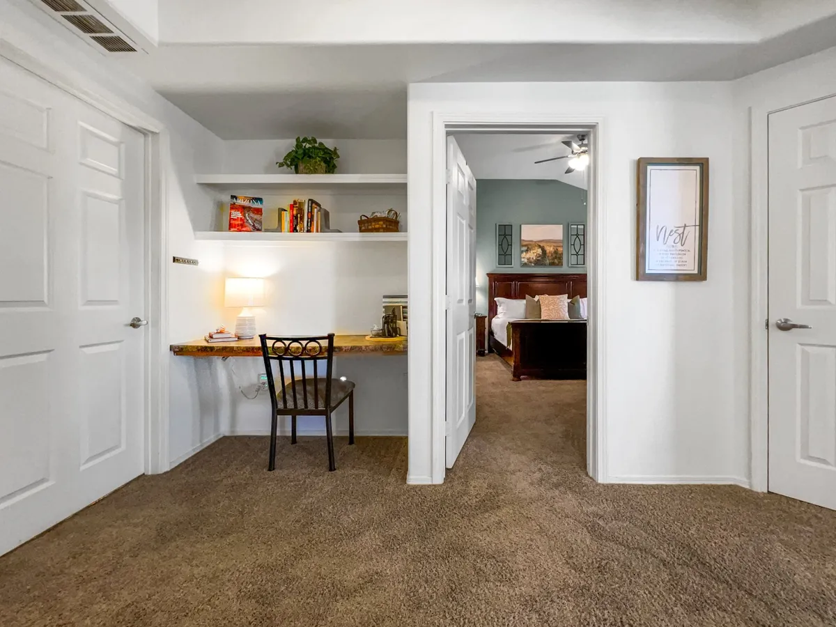 This image features a desk with a chair, shelves adorned with books and decor, and a doorway leading to a bedroom. The area is cozy and functional, evoking the feel of a personal workspace that is perfect for reading, working, or enjoying quiet moments.