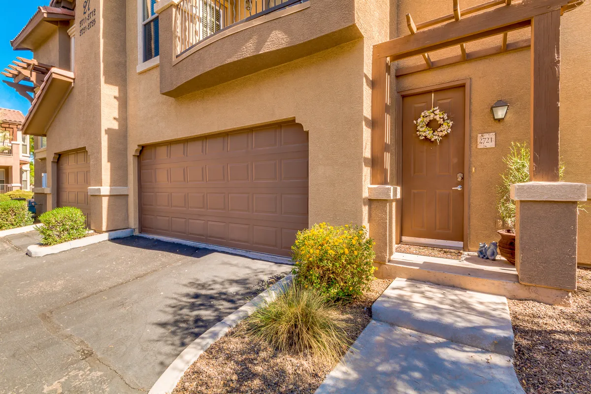 This exterior shot captures the townhome's welcoming front entrance, featuring a decorative wreath on the door, a landscaped area with greenery, and a view of the garage. The porch area suggests a comfortable spot for morning coffee or greeting arriving guests.