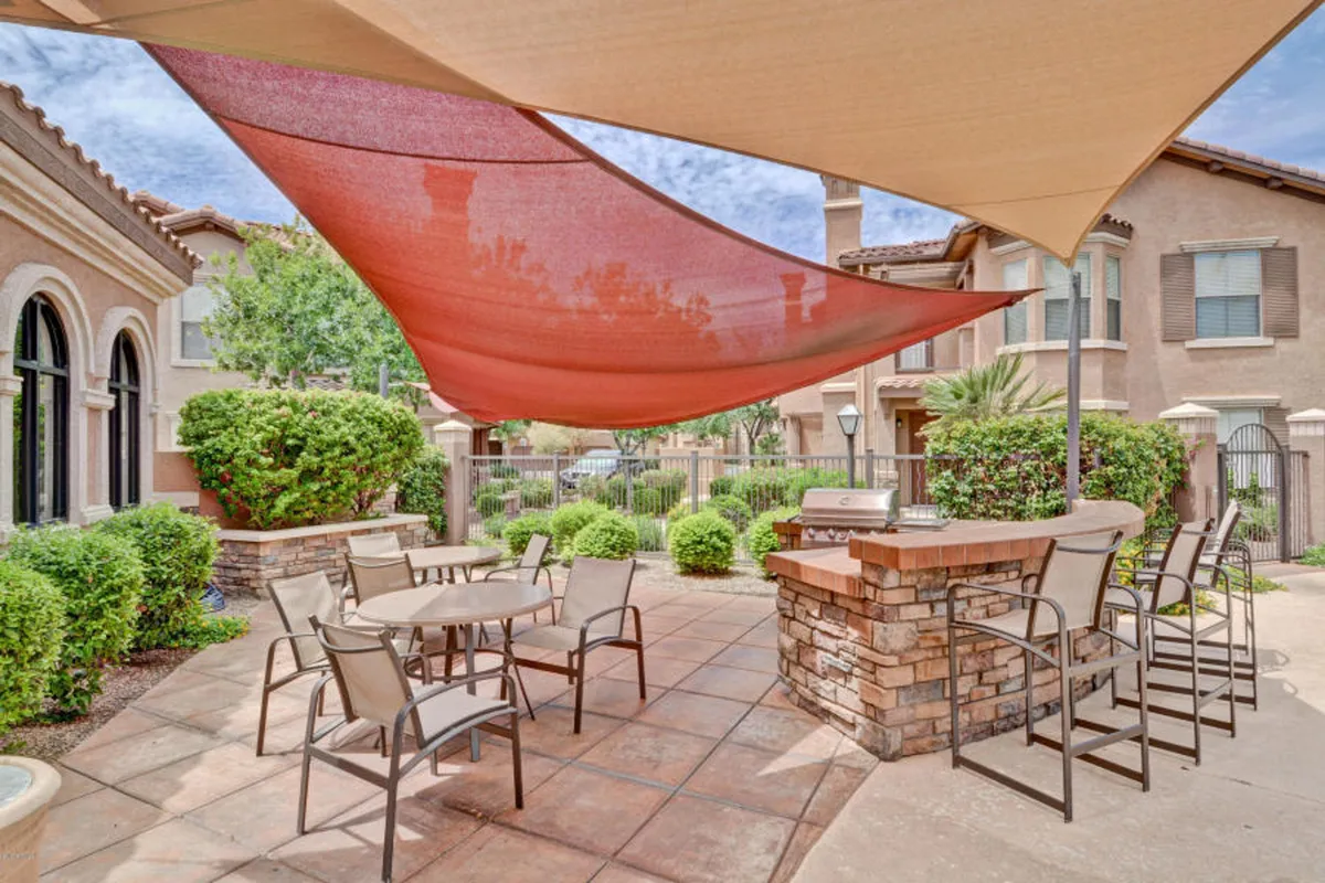 An inviting outdoor seating area complete with a modern grill station under shade sails, surrounded by lush landscaping. The space is designed for social gatherings, barbecues, and relaxed dining, evoking the atmosphere of a casual family get-together in the fresh air.