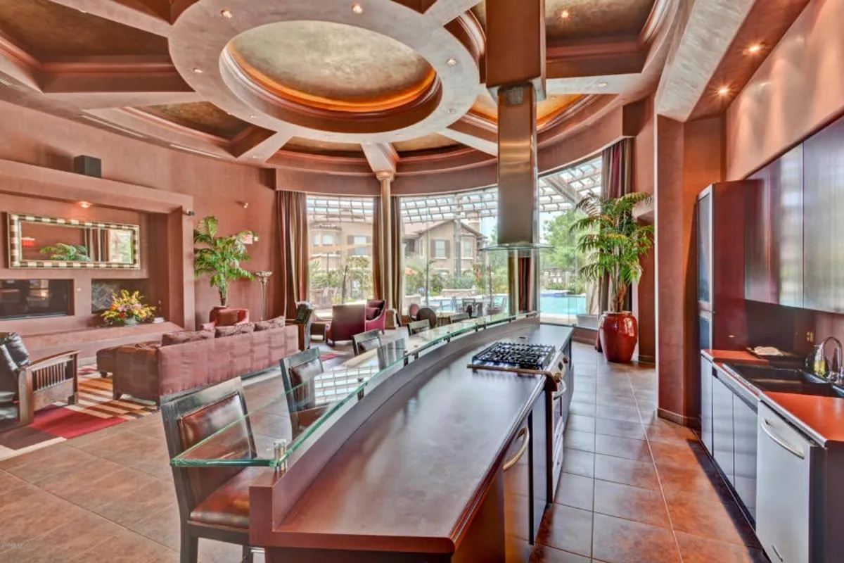 A spacious, elegant interior view reveals a central kitchen island with a stove, curved ceilings, ample seating, and large windows offering glimpses of the pool outside. The layout encourages cooking, entertaining, and social gatherings.