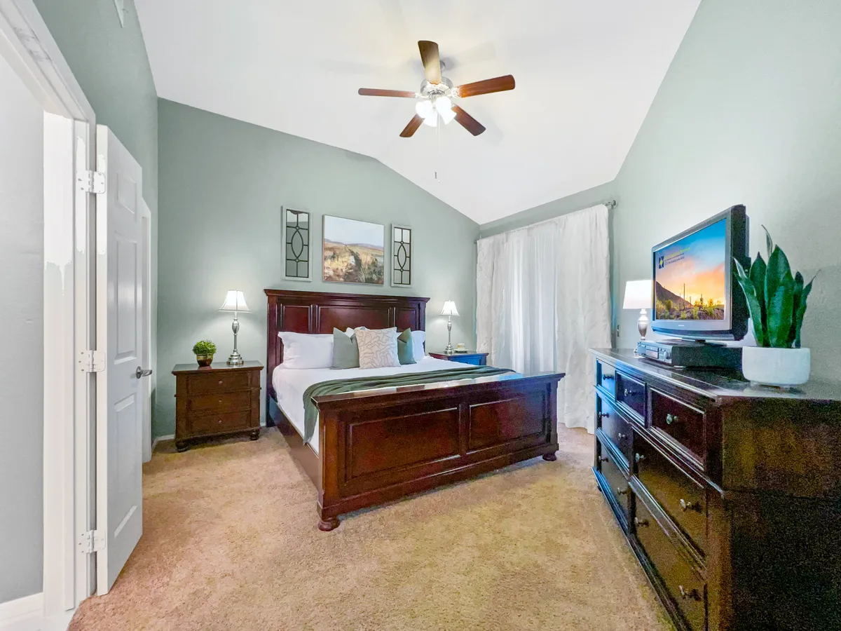 A spacious master bedroom featuring a king-size bed with plush bedding, nightstands on either side, a ceiling fan above, a wall-mounted television, and calming artwork. The room exudes a serene and restful ambiance, ideal for relaxation and privacy.