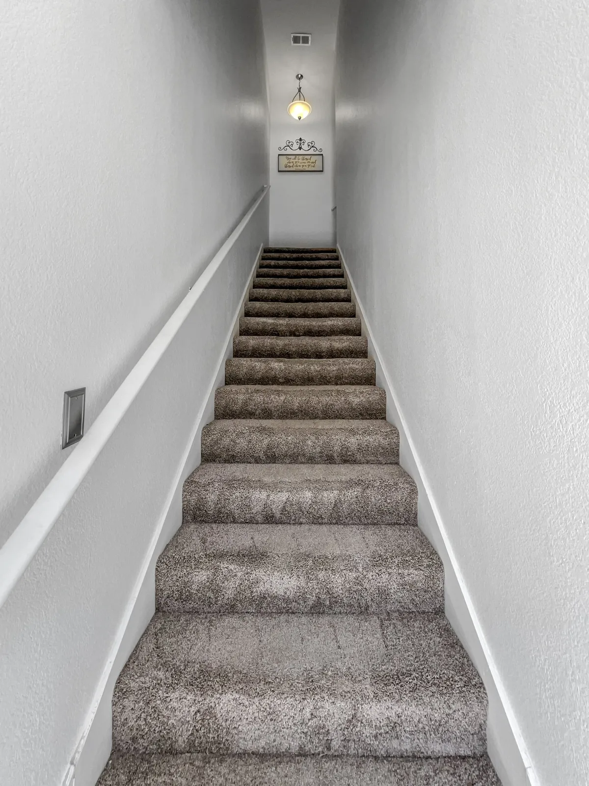 A well-lit staircase with a modern light fixture and a welcoming sign leads up from the entryway, hinting at the transition from the private garage to the main living spaces above.