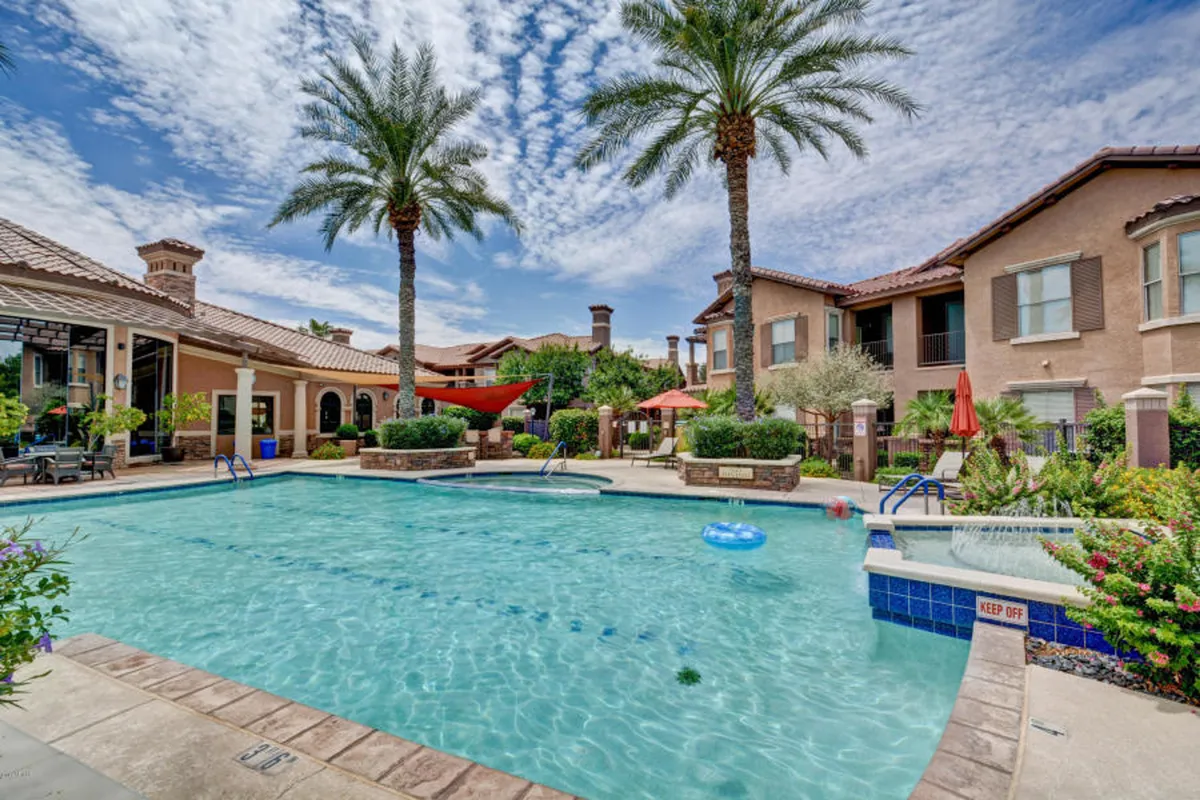 A vibrant exterior view of the community swimming pool, flanked by palm trees and surrounded by lounge areas with colorful umbrellas. The scene is bright and lively, with ample space for swimming, sunbathing, and socializing. The ambiance suggests both relaxation and fun, suitable for all ages.