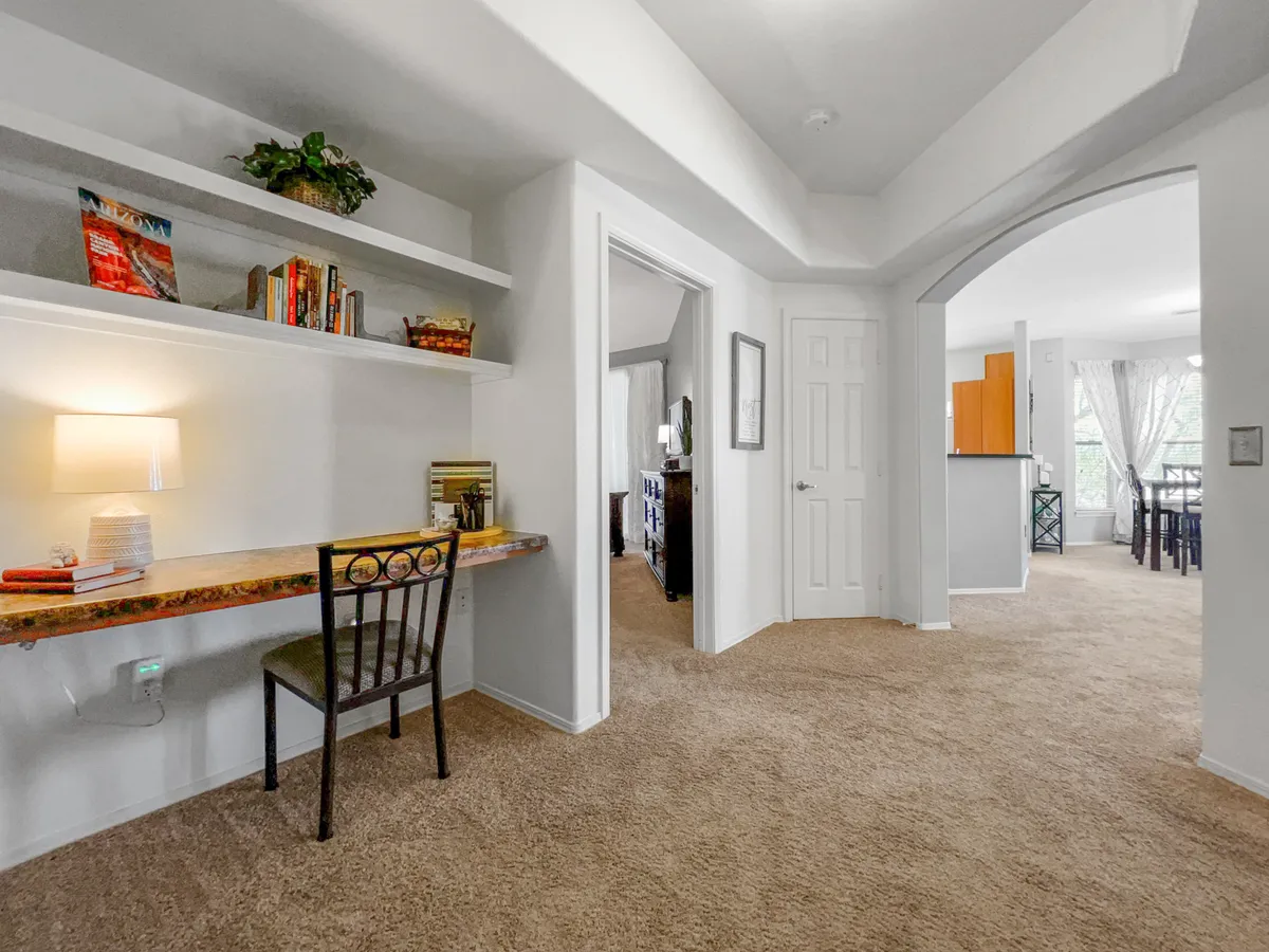 A serene nook featuring a tidy writing desk with a bookshelf, warm lighting, and an archway leading to the dining area. The dedicated workspace is ideal for focused workdays, while the open layout suggests flexibility for work and relaxation.