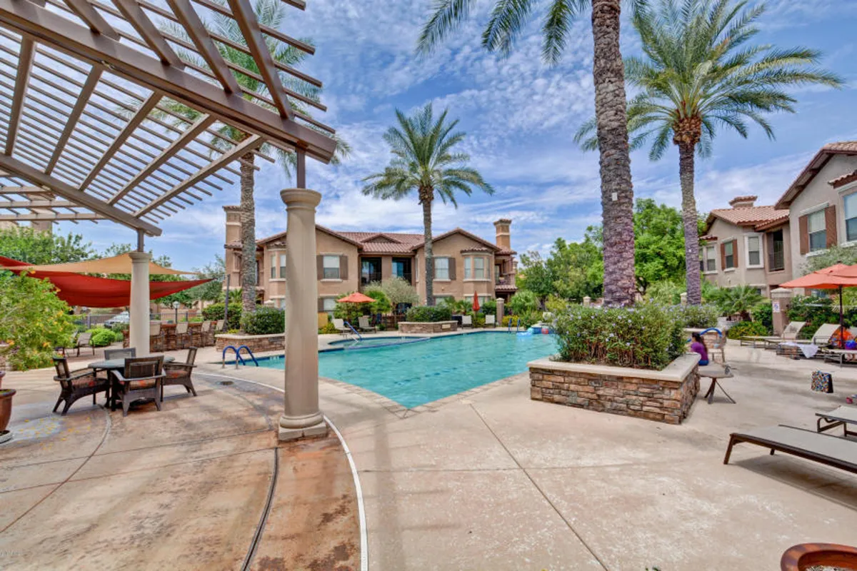 This exterior image features a sparkling swimming pool surrounded by palm trees, inviting lounge chairs, and an outdoor dining area. The scene is sun-soaked, evoking a resort-like atmosphere perfect for relaxation and socializing under Arizona’s clear skies.