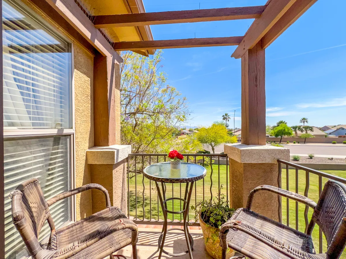 An inviting balcony scene with wicker chairs, a glass table adorned with flowers, and potted plants. The space is surrounded by greenery, emphasizing relaxation and a connection to nature—perfect for quiet mornings or gatherings.